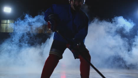 Male-hockey-player-with-a-puck-on-the-ice-arena-shows-dribbling-moving-directly-into-the-camera-and-looking-directly-into-the-camera-against-a-dark-background-in-the-smoke